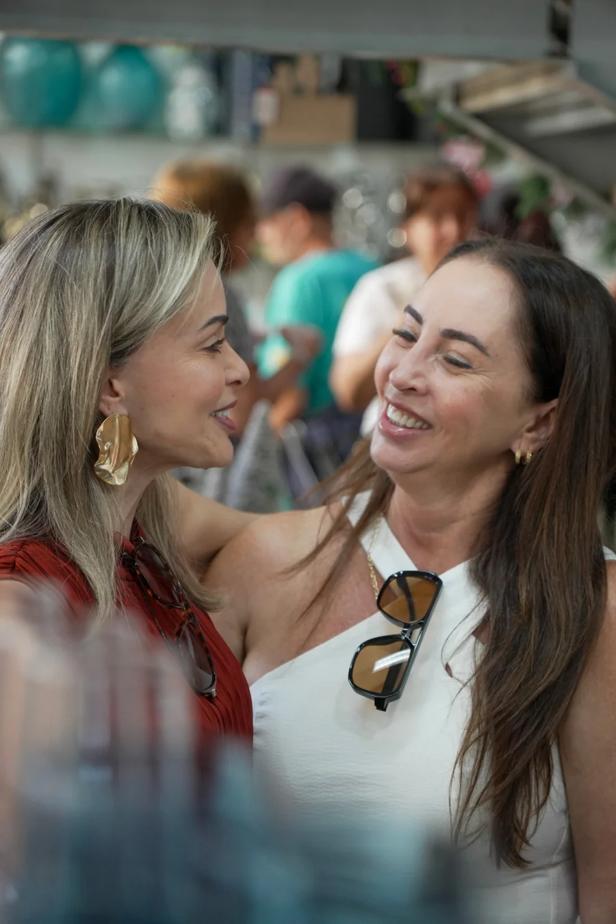 Duas mulheres frente a frente rindo alto dentro da loja, uma de vermelho e a outra de branco, no meio dos convidados