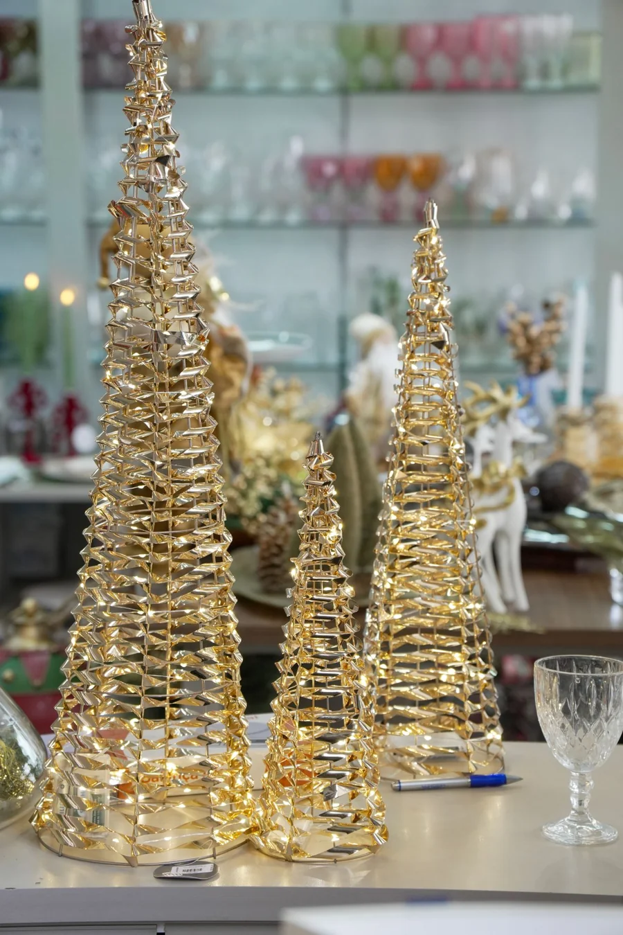 Três árvores de Natal douradas e acesas sobre o balcão da loja, com taças de cristal ao lado e prateleiras de vidro colorido ao fundo