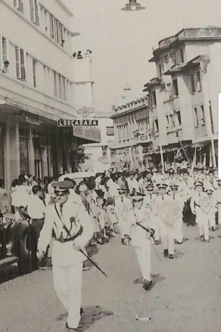 Fotografia antiga de um desfile passando pela rua, com gente na calçada e o letreiro Louça Dada acima da loja