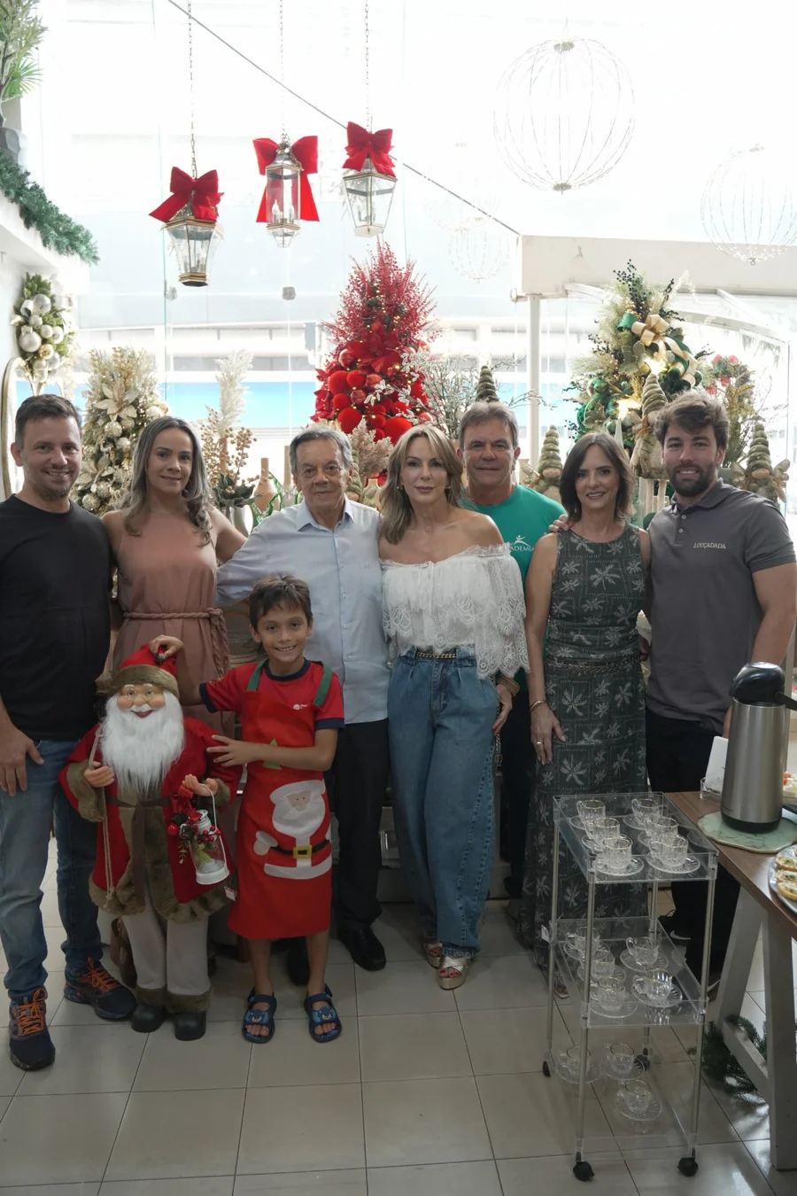 A família da Louçadada reunida no salão da loja no Natal, com uma criança na frente, entre árvores montadas e lanternas de teto