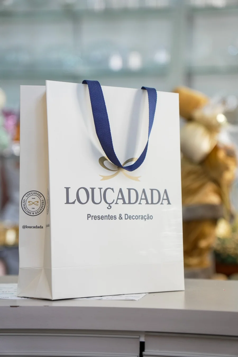 A sacola branca da Louçadada em cima do balcão, com fita azul e o selo da casa impresso na lateral